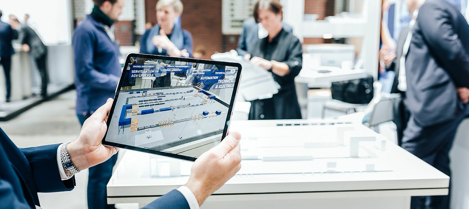 A trade fair visitor is holding a tablet on which a digital factory can be seen.