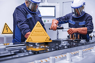 Two Leadec employees in protective clothing repairing a defective battery, in front of them a warning sign for working under high voltage.