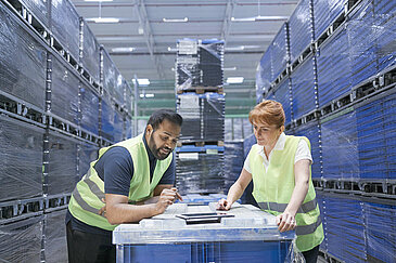 Two Leadec employees with safety vests looking on a tablet between pallets with load carriers in a warehouse.