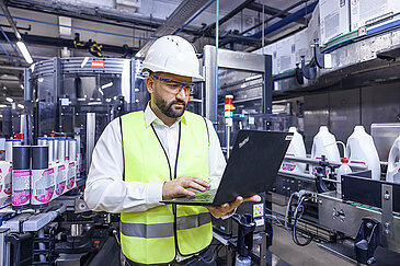 A Leadec employee in safety clothing with a laptop in the middle of conveyor belts with cleaning agents.